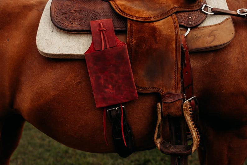 Op de afbeelding: Close-up van een bruin leren zadel met een rode leren tasje bevestigd. Het zadel staat op een bruin paard. De stijgbeugel is zichtbaar. Het zadel heeft een witte pad.