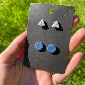 Puede incluir: Dos pares de pendientes de botón se muestran en una tarjeta negra. Los pendientes superiores son triangulares con un diseño gris moteado. Los pendientes inferiores son redondos y azules con purpurina. La tarjeta se sostiene en una mano.