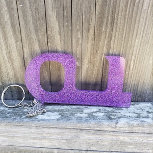 May include: Purple glitter resin keychain in the shape of the letters "OL". The keychain has a silver-colored butterfly charm and a silver key ring. The background is a weathered wooden surface.