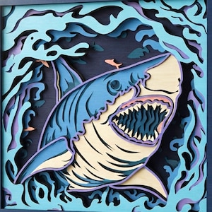 May include: A square multi-layered wall art piece featuring a shark swimming through stylized waves in shades of blue, light blue, and purple. The shark is depicted with an open mouth showing sharp teeth. The text DXF AI SVG is visible.