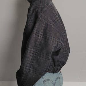 May include: A dark gray plaid jacket with a high collar and a fitted waist. The jacket has a classic plaid pattern in shades of gray and brown. The jacket is paired with light blue denim jeans.