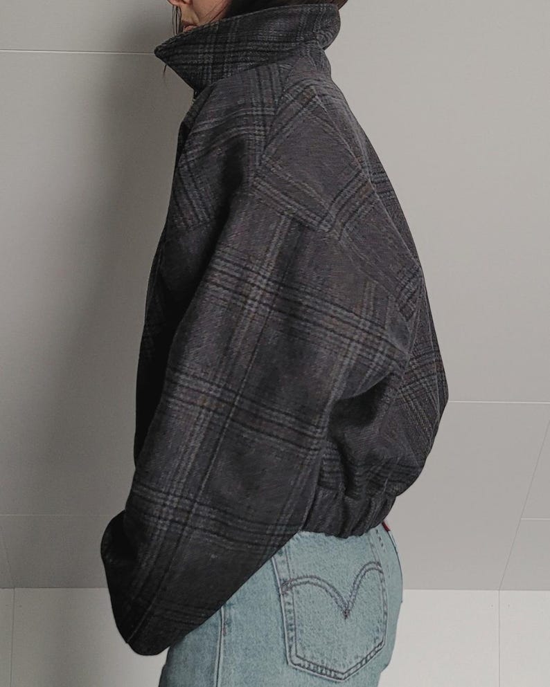 May include: A dark gray plaid jacket with a high collar and a fitted waist. The jacket has a classic plaid pattern in shades of gray and brown. The jacket is paired with light blue denim jeans.