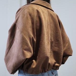 May include: A brown suede jacket with a collared neckline and elastic hem. The jacket has a relaxed fit and long sleeves. The model is wearing blue jeans. The jacket is a neutral color and can be worn with many outfits.