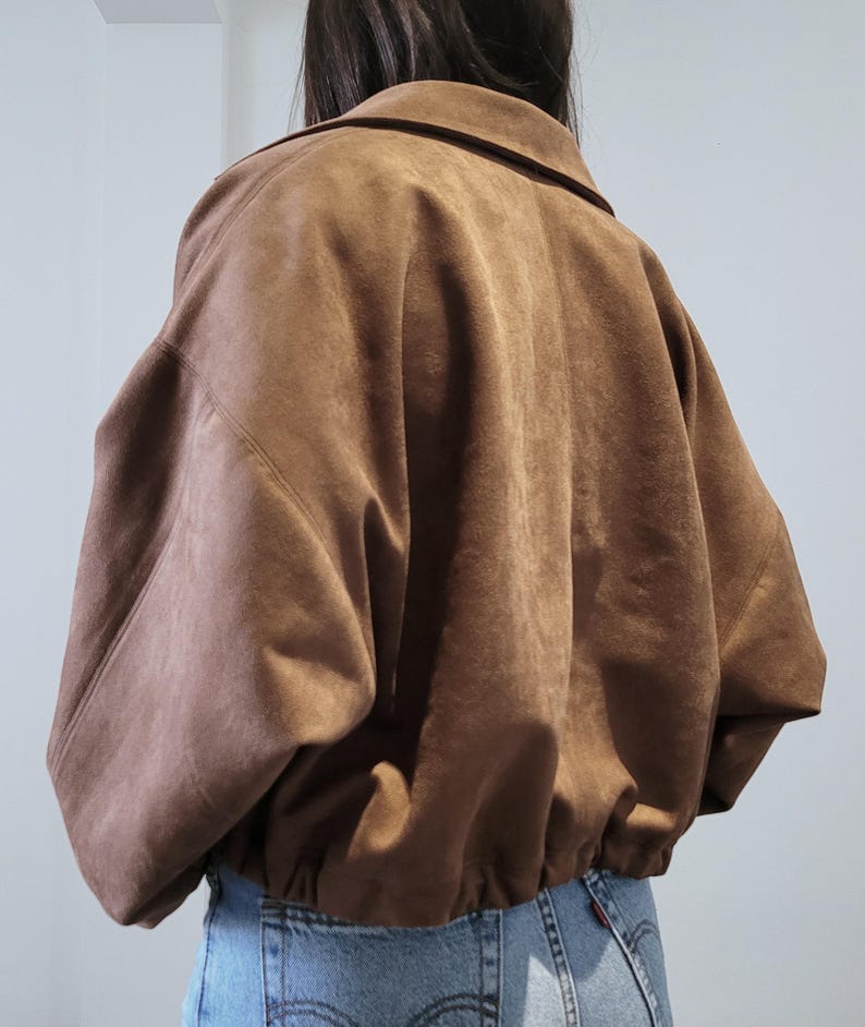 May include: A brown suede jacket with a collared neckline and elastic hem. The jacket has a relaxed fit and long sleeves. The model is wearing blue jeans. The jacket is a neutral color and can be worn with many outfits.