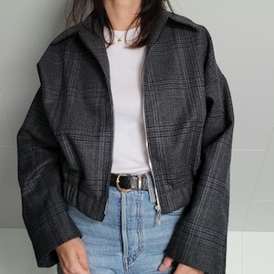 May include: A gray plaid jacket with a zipper closure. The jacket has a collar and long sleeves. The model is wearing a white t-shirt, blue jeans, and a black belt with a gold buckle.