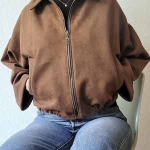 May include: A brown suede jacket with a zipper closure and a collared neckline. The jacket has a relaxed fit and is paired with blue jeans. The jacket's color is a warm, earthy tone, and the zipper is a contrasting metallic color.
