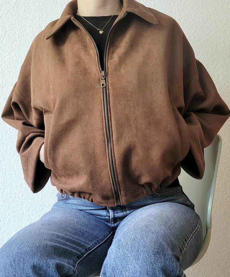 May include: A brown suede jacket with a zipper closure and a collared neckline. The jacket has a relaxed fit and is paired with blue jeans. The jacket's color is a warm, earthy tone, and the zipper is a contrasting metallic color.