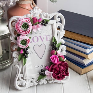 Puede incluir: Marco de fotos blanco adornado con un arreglo floral rosa y burdeos. El marco tiene un paspartú blanco con la palabra "LOVE" y un corazón rosa en el centro. El marco mide 10x15cm.