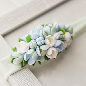 Op de afbeelding: Een lichtgroen hoofdband met een cluster van witte en lichtblauwe viltbloemen. De bloemen zijn in een cirkelvormige vorm gerangschikt en hebben groene viltbladeren.