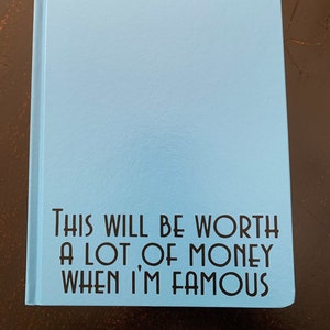 May include: A light blue notebook with the text "This will be worth a lot of money when I'm famous" printed on the front.