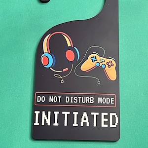 May include: Black door hanger with a gamer theme. It features a pair of headphones, a video game controller, and the text "DO NOT DISTURB MODE INITIATED" in a pixelated font.