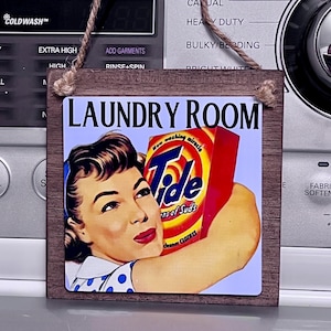 May include: A rectangular wooden sign with a vintage-style image of a woman holding a Tide detergent box. The sign reads "LAUNDRY ROOM" in bold letters and has a twine loop for hanging.