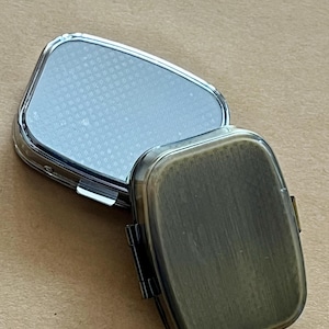 May include: An open, rectangular pill box with a silver-colored lid and a bronze-colored base. The lid is reflective and has a textured pattern. The base has a hinged closure. The pill box is designed to hold medication or small items.