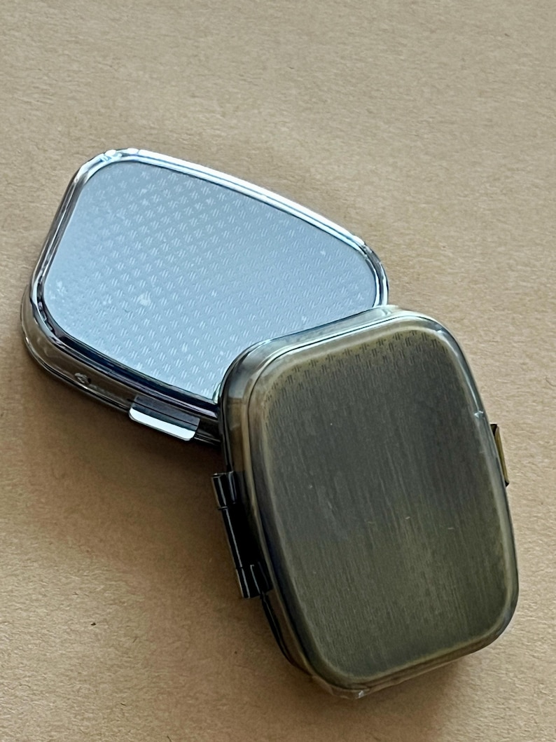 May include: An open, rectangular pill box with a silver-colored lid and a bronze-colored base. The lid is reflective and has a textured pattern. The base has a hinged closure. The pill box is designed to hold medication or small items.