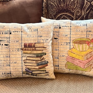 May include: Two square throw pillows featuring a vintage library checkout card print on a beige background. One pillow displays a stack of books, while the other shows a teacup resting on three books. Both include printed dates and text.