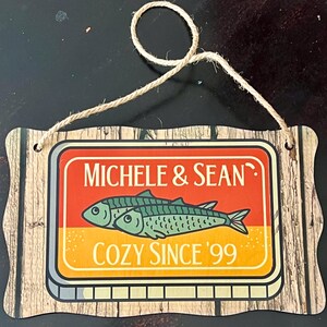 May include: Wooden sign with a rope hanger. The sign features the text "MICHELE & SEAN" above two green fish and the text "COZY SINCE '99" in a retro design. The background has a wood grain pattern.