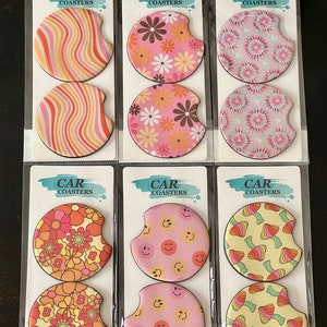 May include: Six sets of car coasters with different designs. Each set includes two round coasters. The designs include pink and orange swirls, pink and brown flowers, pink and blue tie-dye, orange and pink flowers, pink and orange smiley faces, and yellow and orange mushrooms.