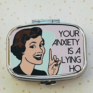 May include: A silver-colored pill box with a retro illustration of a woman pointing her finger. The text on the box reads "YOUR ANXIETY IS A LYING HO". The background is a light pink and blue.