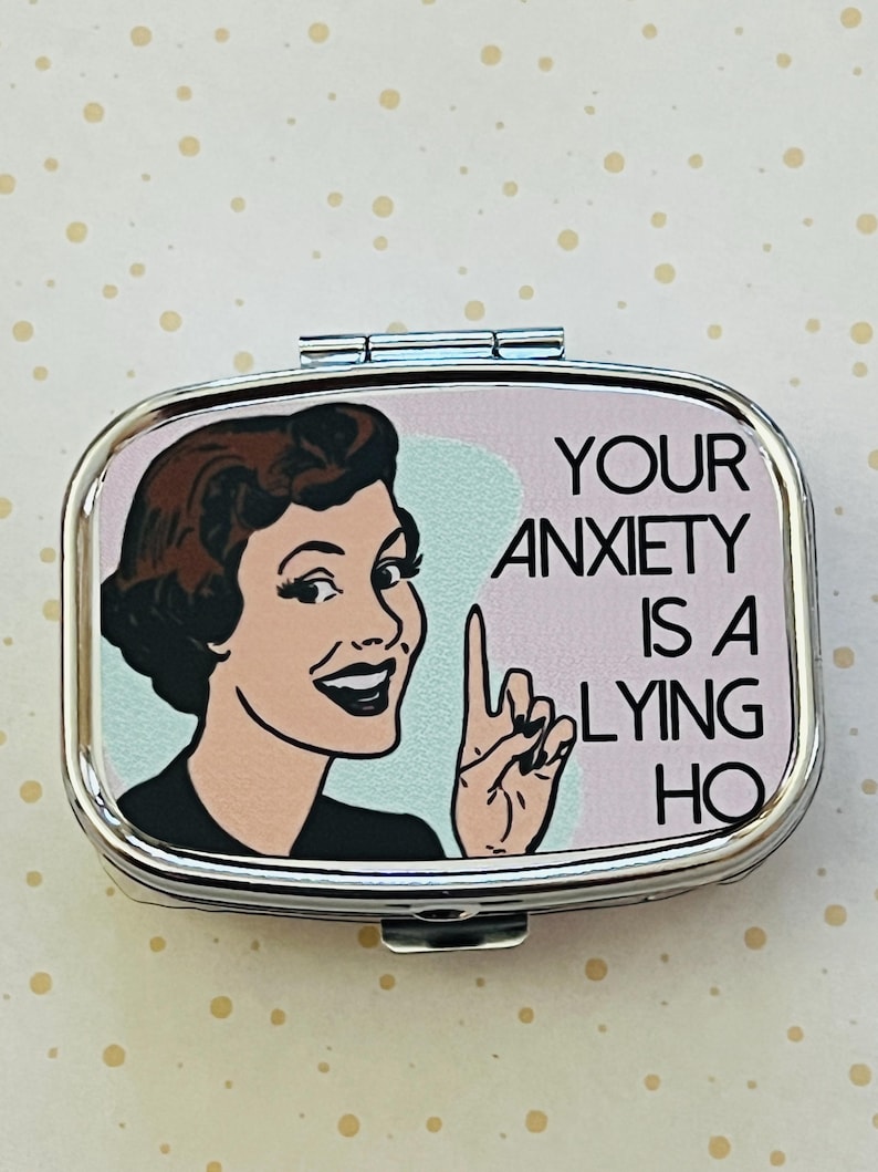 May include: A silver-colored pill box with a retro illustration of a woman pointing her finger. The text on the box reads "YOUR ANXIETY IS A LYING HO". The background is a light pink and blue.