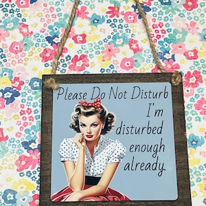 May include: A square wooden sign with a jute rope hanger featuring a light blue background, a retro-style illustration of a person with a red hair bow and polka dot shirt, and the text: Please Do Not Disturb I'm disturbed enough already.