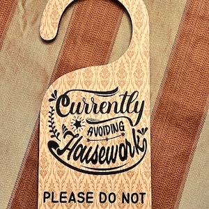 May include: A wooden door hanger with a beige and brown damask pattern. The hanger has a cutout for a door knob and features the text "Currently Avoiding Housework" in black cursive lettering with a star and arrow. The bottom of the hanger reads "Please Do Not Disturb" in black text.
