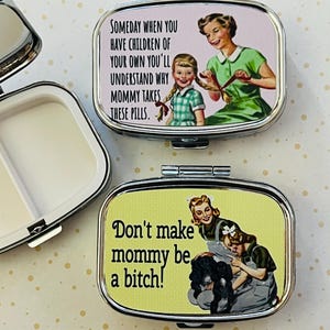 May include: Two silver pill cases are open, revealing white interiors. The cases feature retro-style images and text. One reads "Someday when you have children..." and the other says "Don't make mommy be a bitch!"