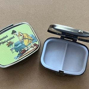 May include: Two rectangular pill boxes with a hinged lid. One is open, revealing a white interior divided into two sections. The closed box has a green background with the words "Medicated & Motivated" and a vintage illustration of a woman.