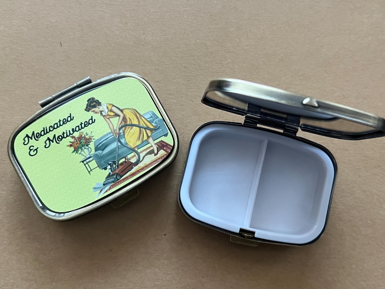 May include: Two rectangular pill boxes with a hinged lid. One is open, revealing a white interior divided into two sections. The closed box has a green background with the words "Medicated & Motivated" and a vintage illustration of a woman.