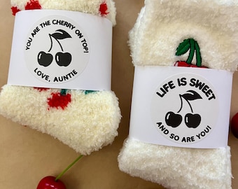 Op de afbeelding: Een paar crèmekleurige, pluizige sokken met rode kersenpatronen. Elke sok is omwikkeld met een witte papieren band. Op de ene band staat "YOU ARE THE CHERRY ON TOP! LOVE, AUNTIE" en op de andere "LIFE IS SWEET AND SO ARE YOU!" met kersafbeeldingen.