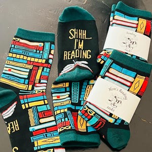 May include: Three black socks with teal cuffs and heels featuring a colorful pattern of stacked books. One sock displays the text SHHH... I'M READING above an open book graphic. Two socks are folded and wrapped in white paper bands labeled Reading Socks.