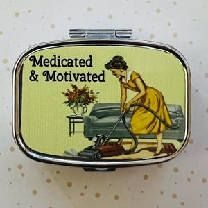 May include: Silver pill box with a retro illustration. The image features a woman in a yellow dress vacuuming a living room. Text on the box reads "Medicated & Motivated". The background is a light green color with a sofa, table, and flowers.