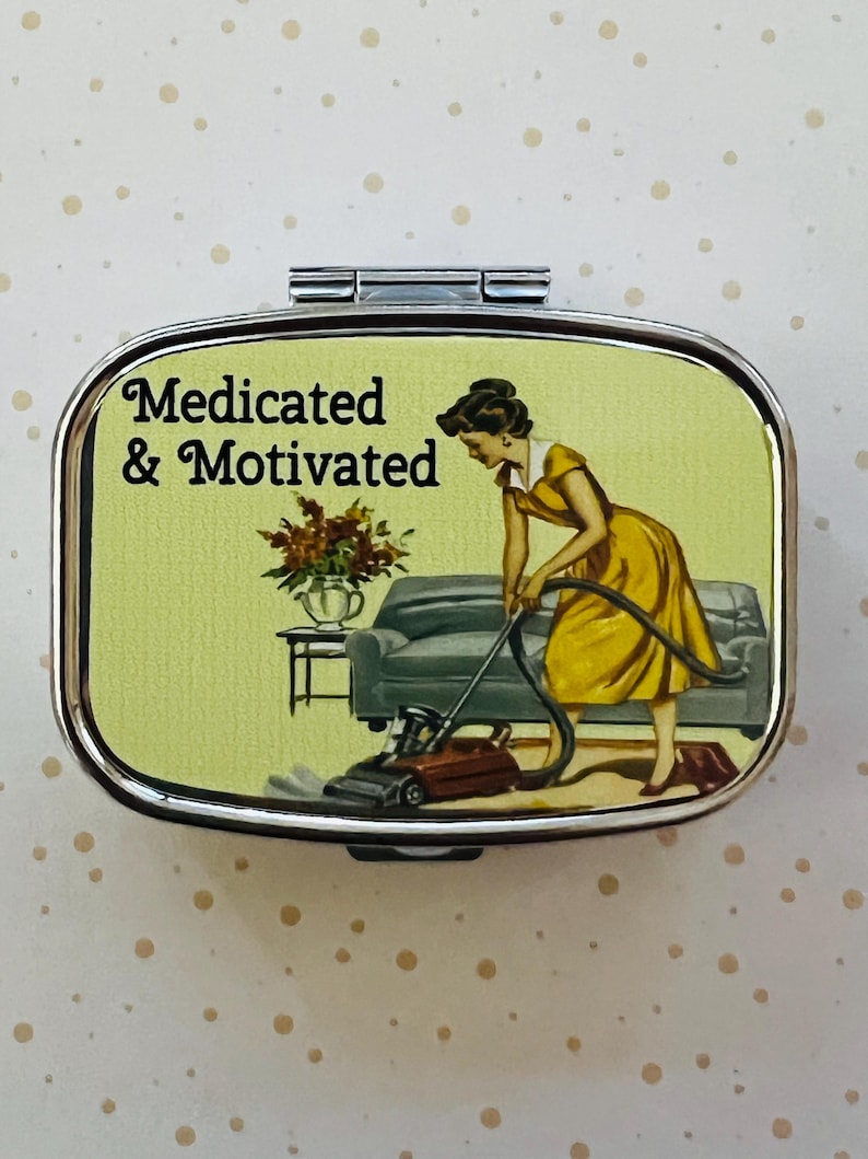 May include: Silver pill box with a retro illustration. The image features a woman in a yellow dress vacuuming a living room. Text on the box reads "Medicated & Motivated". The background is a light green color with a sofa, table, and flowers.