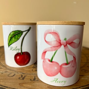 May include: Two white ceramic jars with wooden lids, each featuring a cherry design. The jar on the left has a red cherry with a green leaf and the name "Riley" written below. The jar on the right has two pink cherries with a pink bow and the name "Avery" written below.