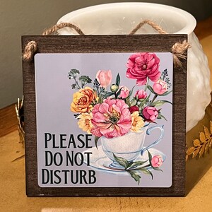 May include: A square wooden sign with a floral design and the text "PLEASE DO NOT DISTURB." The artwork features a teacup overflowing with pink, yellow, and red flowers. The sign has a brown frame and a jute rope for hanging.