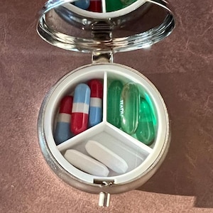 May include: A silver, round pill case with a hinged lid and a built-in mirror. The case is open, revealing three compartments filled with pills of various colors and shapes. The pills include red, blue, and white capsules.
