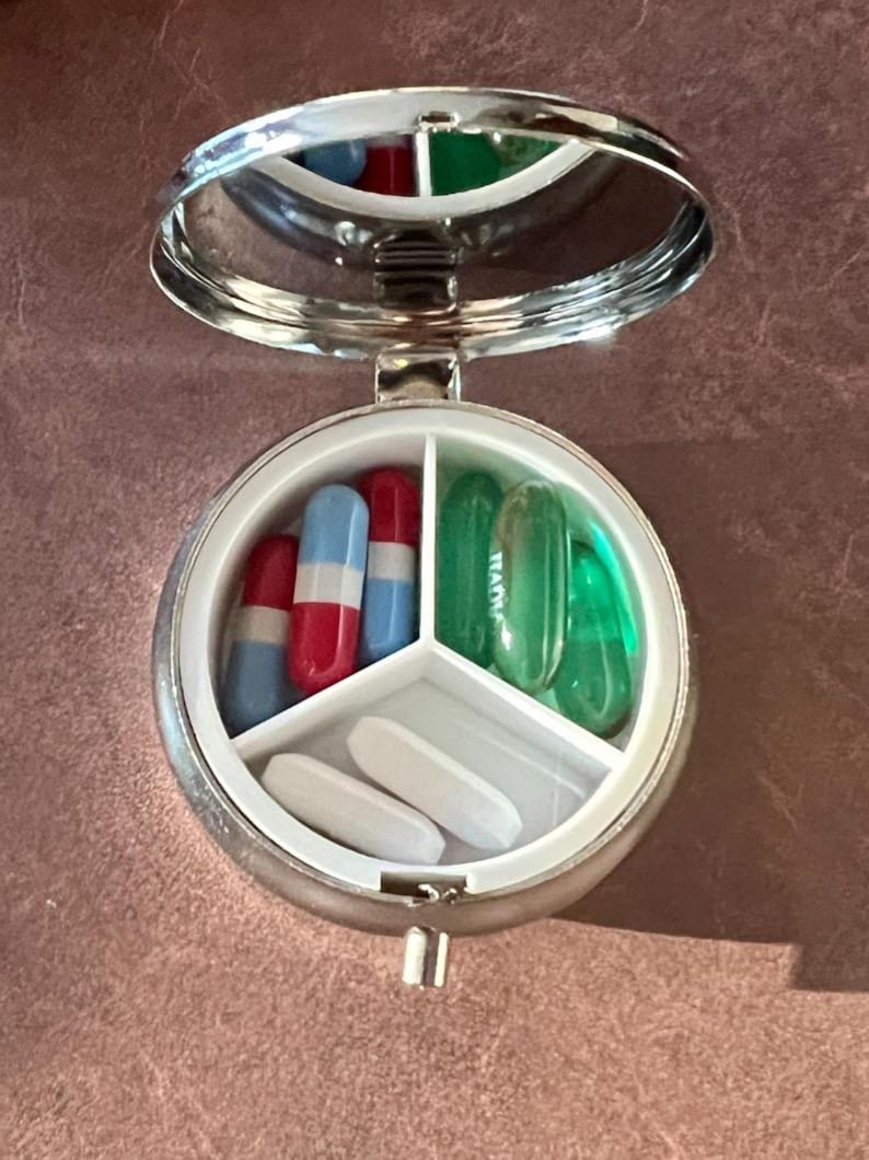 May include: A silver, round pill case with a hinged lid and a built-in mirror. The case is open, revealing three compartments filled with pills of various colors and shapes. The pills include red, blue, and white capsules.