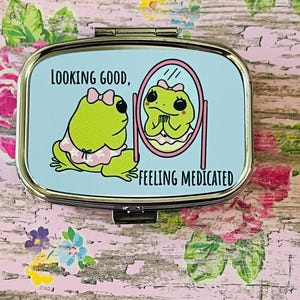 May include: A rectangular metal pill case with a light blue top featuring a cartoon green frog wearing a pink bow and white patterned top, looking into a pink oval mirror. The text reads LOOKING GOOD, FEELING MEDICATED in black lettering.