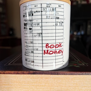 May include: White ceramic mug with a black and white library card design. The mug has a bamboo lid and the words "Book Money" are written in red on the front.