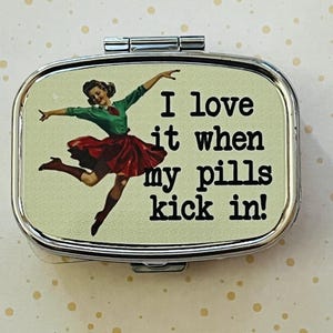 May include: A silver metal pill box with a hinged lid. The box features a vintage-style image of a dancing woman in a green top and red skirt, with the text "I love it when my pills kick in!"