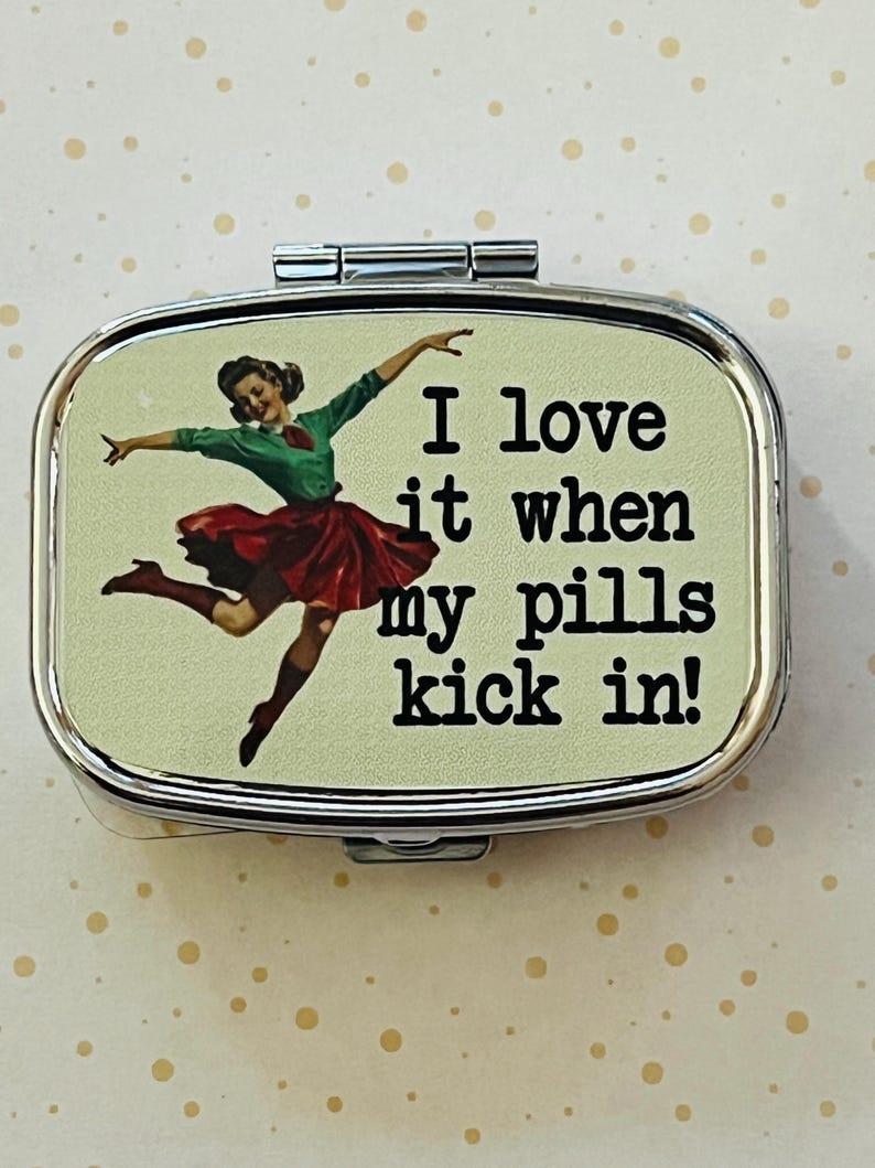 May include: A silver metal pill box with a hinged lid. The box features a vintage-style image of a dancing woman in a green top and red skirt, with the text "I love it when my pills kick in!"