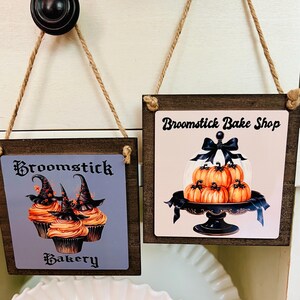 May include: Two square wooden hanging signs with jute rope. The left sign features orange cupcakes with black witch hats and the text Broomstick Bakery. The right sign shows orange pumpkins on a black cake stand with a glass dome and the text Broomstick Bake Shop.