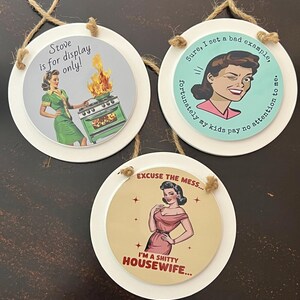 May include: Three round white hanging signs with jute twine. One features a woman by a flaming stove with text "Stove is for display only!". Another shows a winking woman with "Sure, I set a bad example, fortunately my kids pay no attention to me.". The third says "Excuse the mess... I'm a shitty housewife..." with a woman.