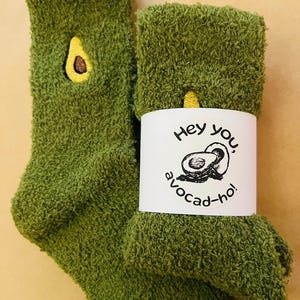 May include: A pair of green fuzzy socks with an embroidered avocado design. One sock features a yellow avocado pit. A white label wrapped around the socks reads "Hey you, avocado-ho!" with a black avocado illustration.