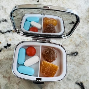 May include: A small, silver pill case with a mirror lid, open to reveal a variety of pills and gummies. The case is divided into two compartments, one with blue and white pills, and a red pill. The other compartment contains brown and orange gummies.