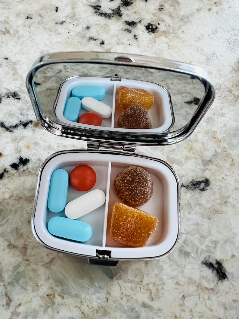 May include: A small, silver pill case with a mirror lid, open to reveal a variety of pills and gummies. The case is divided into two compartments, one with blue and white pills, and a red pill. The other compartment contains brown and orange gummies.
