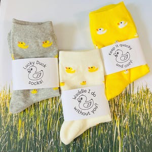 May include: Three pairs of crew socks in gray, cream, and yellow, each featuring a repeating pattern of small yellow rubber ducks. Each pair is secured with a white paper band displaying a duck illustration and text: Lucky Duck socks!, Waddle I do without you?, and Keep it quacky and cozy.