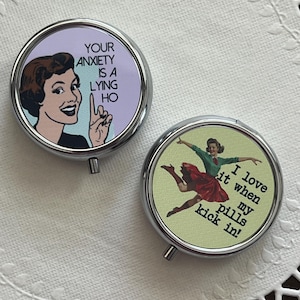 May include: Two round, silver pill cases. One features a vintage illustration with the text "YOUR ANXIETY IS A LYING HO." The other has a woman dancing with the text "I love it when my pills kick in!" on a green background.