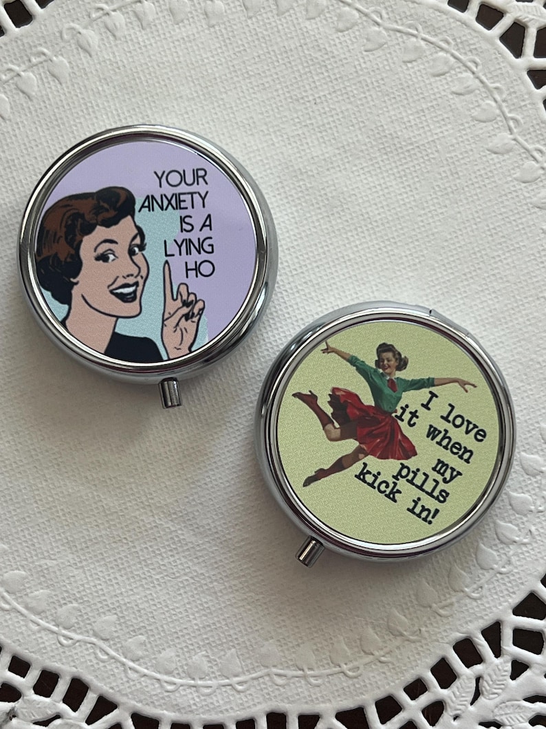 May include: Two round, silver pill cases. One features a vintage illustration with the text "YOUR ANXIETY IS A LYING HO." The other has a woman dancing with the text "I love it when my pills kick in!" on a green background.