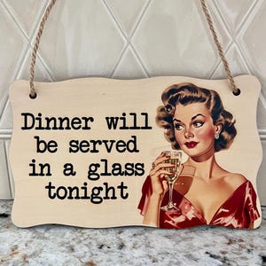 May include: Wooden sign with the text "Dinner will be served in a glass tonight." The sign features a vintage illustration of a woman in a red dress holding a glass of wine. The sign is hung with a natural twine.