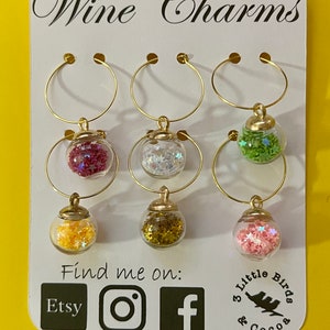 May include: Six gold wine glass charms with different colored glitter inside. The charms are displayed on a white card with the text "Wine Charms" and "Find me on: Etsy Instagram Facebook" and a logo for "3 Little Birds & Cocoa".
